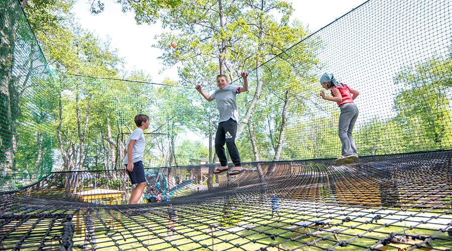 Moments de joie sur un filet de trampoline en plein air Cette image montre des enfants s'amusant sur un filet de trampoline dans un parc d'accrobranche, capturant l'énergie et la joie des activités en plein air entourées de nature verdoyante.